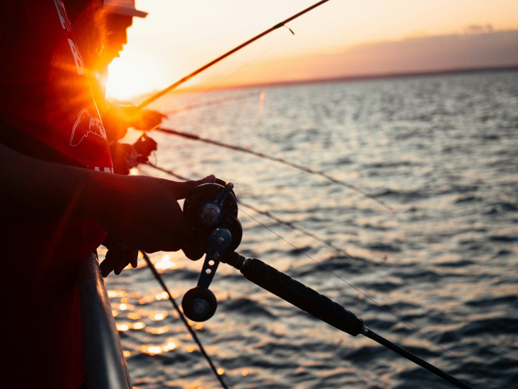 An angler's hands on a rod and reel, silhouetted against a low orange sun over the sea